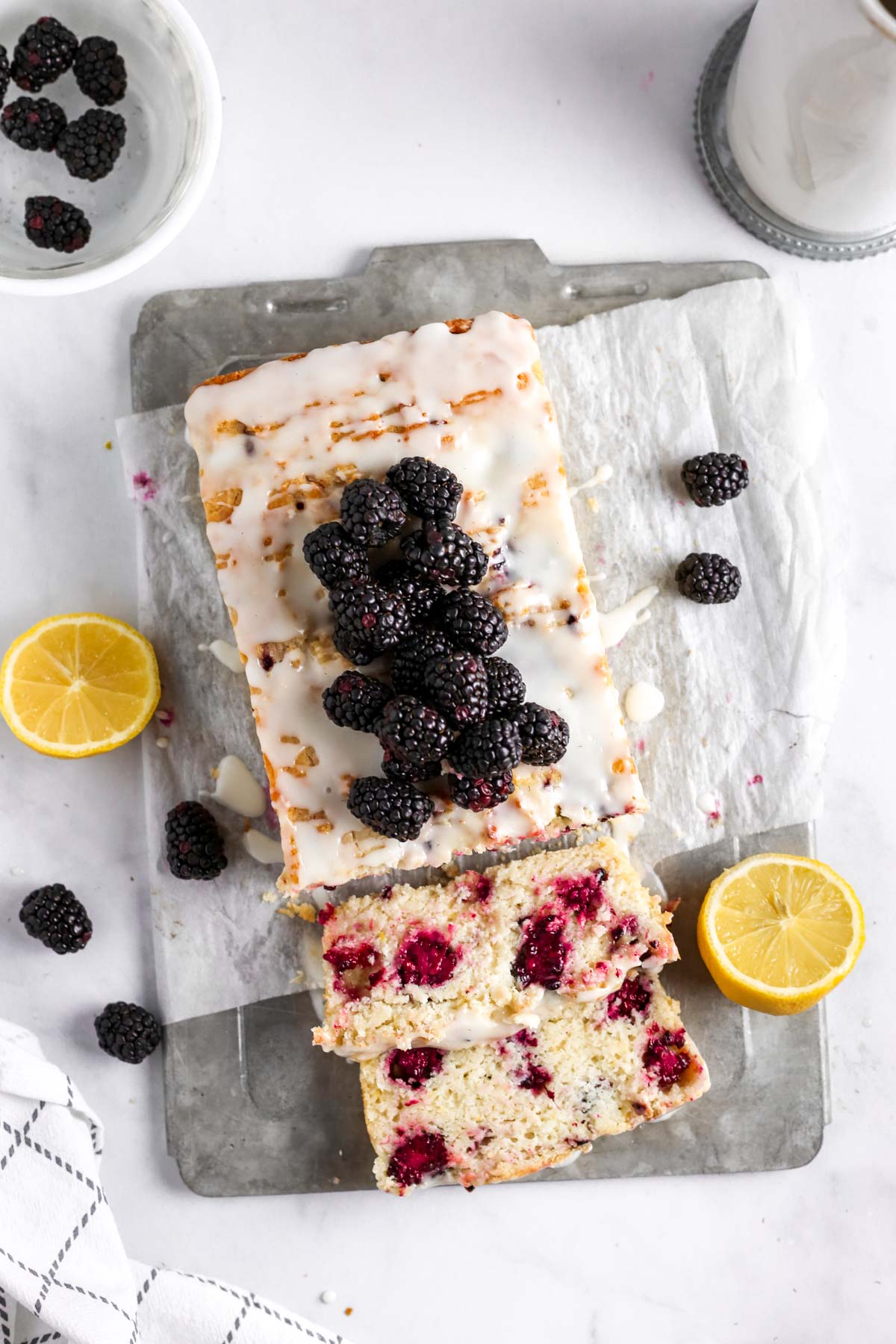 a blackberry lemon loaf cake on a gray tray with parchment paper. The cake has fresh blackberries and lemon zest on top. It is sliced, showing the blackberries inside the lemon cake.