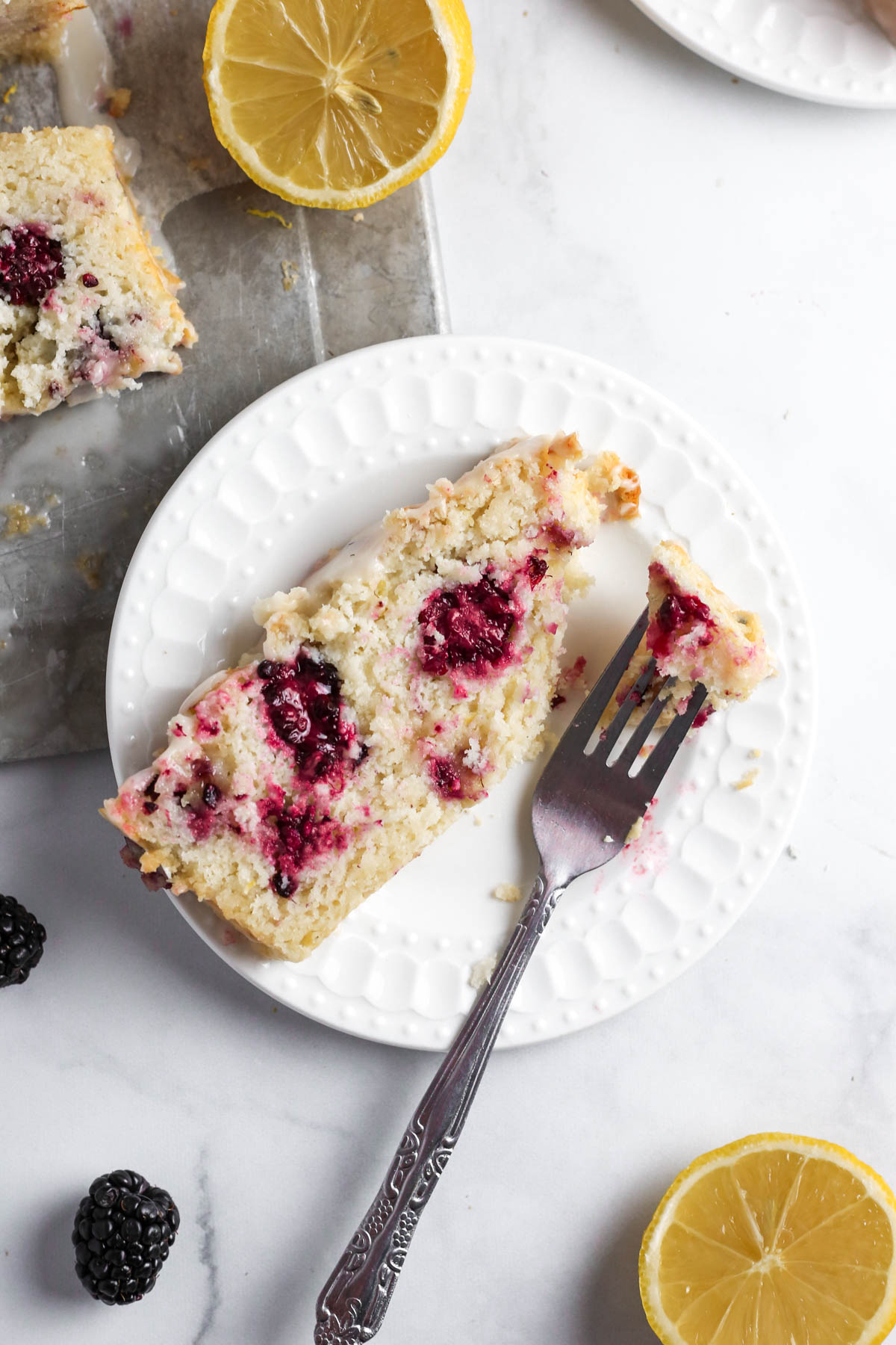 a slice of blackberry lemon bread with lots of blackberries baked inside. The slice is on a white dessert plate with a fork.