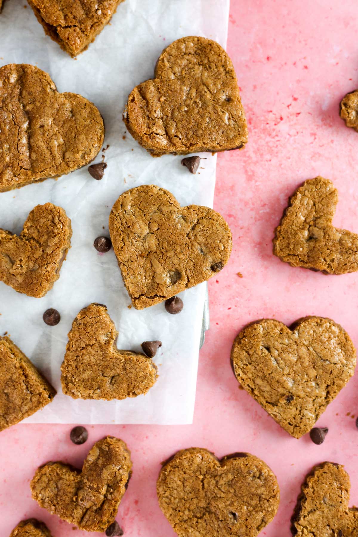 multiple heart shaped chocolate chip cookies resting on parchment paper on a pink surface.