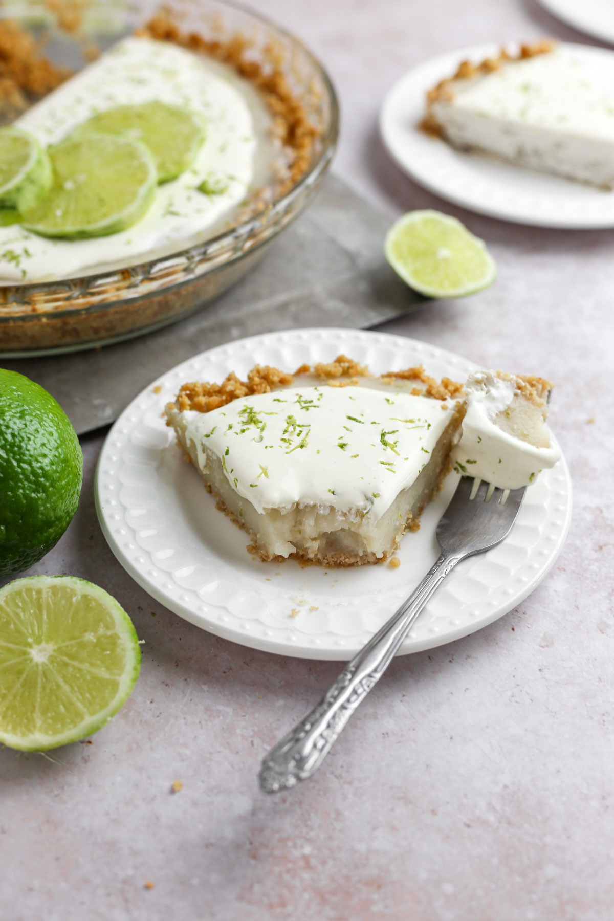 slice of key lime pie with coconut whipped cream and fresh lime zest on top. There is a fork next to the slice, and fresh limes around the photo.